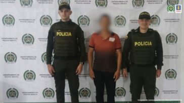 En la imagen se visualiza al capturado de espalda junto a dos uniformados de la Policía Nacional. En la parte posterior se observa el banner que identifica a la Policía Nacional del departamento de Arauca.