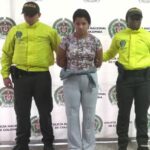 En la fotografía está la procesada con una camiseta, una sudadera gris y un saco amarrado a la cintura con dos servidores de la Policía Nacional que usan chaqueta verde fluorescente y al fondo hay un pendón de la Policía con sus logos en mosaico.