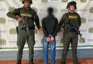 En la imagen se observa al investigado de espaldas junto a dos uniformados de la Policía Nacional. Detrás de ellos el banner que identifica al Departamento de Policía Arauca