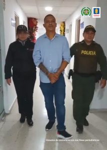 En la fotografía se aprecia en acción de caminata por un pasillo a Jaime Duván Dávalos Giraldo. Lleva los brazos adelante y esposado. Viste camisa azul de mangas recogidas hasta los codos; jean azul y zapatillas oscuras. Al lado derecho de él hay una servidora del CTI y al lado izquierdo un uniformado de la Policía Nacional.