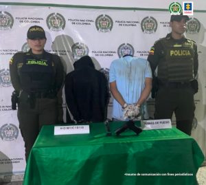 Se observan dos detenidos de espaldas y a cada costado de ellos, dos funcionarios de Policía Nacional.  Adelante una mesa con el arma incautada y atrás un pendón de Policía Nacional