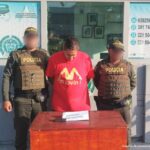 En la imagen está un hombre de pie, con la cabeza agachada, vestido con  una camiseta roja, custodiado por dos uniformados de la Policía Nacional.