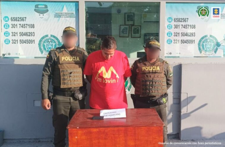 En la imagen está un hombre de pie, con la cabeza agachada, vestido con  una camiseta roja, custodiado por dos uniformados de la Policía Nacional.