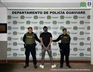 capturado de espaldas esposado custodiado por dos policías. Detrás banner de la Policía Nacional.