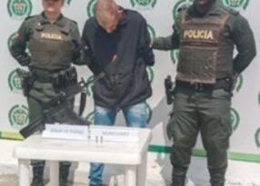 En la imagen se observa el hombre junto a dos uniformados de la Policía Nacional. Frente a ellos una mesa donde se ubico el arma y munición incautada. En la parte posterior se encuentra el banner que identifica a la Policía Nacional.