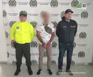 en la imagen se ve una persona detenida bajo custodia de un Policía y un investigador del CTI de la Fiscalía. Detrás suyo un backing institucional.