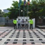 En la fotografía están tres hombres de pie, moreno, gruesos, vestidos con camisetas blancas y bermudas azules, custodiados por dos uniformados de la Policía Nacional. Delante de ellos se ven la droga incautada en paquetes rectangulares.