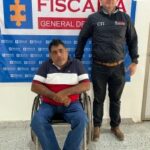 En la fotografía está un hombre sentado en una silla de ruedas, vestido con camiseta de rayas azul oscura, blanca y roja, jean azul y zapatos beige. Se encuentra custodiado por un uniformado del Cuerpo Técnico de Investigación, detrás de ellos hay un pendón de la Fiscalía.