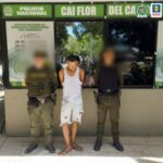 En la imagen está un hombre de pie con la cabeza agachada y los brazos atrás, vestido de camisilla blanca, bermuda de jean azul y sandalias café, custodiado por dos agentes de la Policía Nacional.