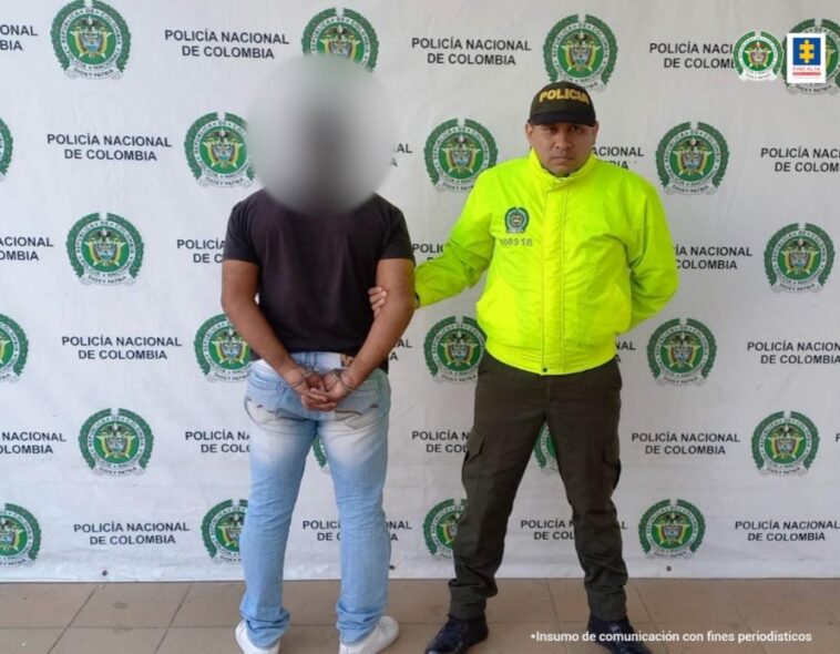 En la imagen está un hombre de pie, de espalda vestido con una camiseta negra y un jean azul custodiado por un uniformado de la Policía Nacional.