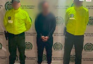 En la fotografía se encuentra un hombre de pie, con las manos esposadas, rostro difuso, vestido de buzo, sudadera y tenis negros custodiado por dos uniformados de la Policía Nacional.