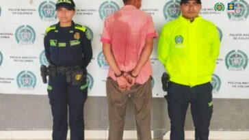 En la fotografía aparece un hombre alto, de pie, de espalda, con las manos esposadas, vestido con una camisa roja manga corta y un pantalón café. Se encuentra custodiado por dos uniformados de la Policía Nacional con un pendón de esa institución.