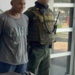 En la imagen aparece un hombre de pie, de estatura baja, moreno, cabellos cortos, vestido de camiseta gris y jean azul, acompañado por un agente de la Policía Nacional. Delante de ellos está el arma incautada.