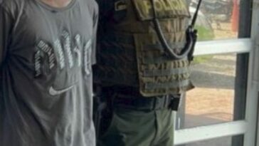 En la imagen aparece un hombre de pie, de estatura baja, moreno, cabellos cortos, vestido de camiseta gris y jean azul, acompañado por un agente de la Policía Nacional. Delante de ellos está el arma incautada.
