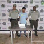 En la fotografía esta un hombre de pie con los brazos atrás, vestido de camiseta azul clara, jean azul, tenis negro, custodiado por dos uniformados de la Policía Nacional. Delante de ellos hay una mesa con la cadena recuperada.