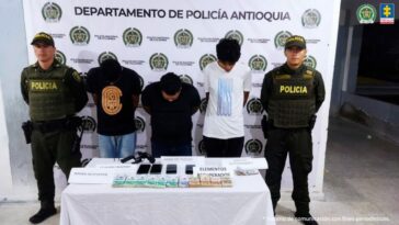 En la imagen aparecen los 3 procesados en medio de dos policía y al frente de ellos hay una mesa con fajos de billetes y un arma tipo revólver.