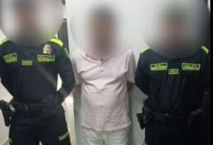 En la foto está un hombre con el rostro tapado, de pie, con los brazos atrás, vestido de camisa rosada, bermuda blanca y medias negras custodiado por dos agentes de la Policía Nacional.