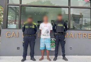 En la imagen se ve a un hombre de pie, de piel oscura, con el rostro desdibujado, vestido con camiseta blanca con short azul y sandalias negras, custodiado por dos agentes de la Policía Nacional en un CAI que señala Daniel Lemaitre.