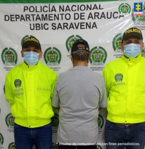 Se visualiza al capturado de espaldas junto a dos uniformados de la Policía Nacional. Detrás el banner que identifica a la Policía Nacional en Saravena (Arauca)