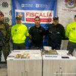 Se observa al procesado junto a un investigador del CTI y uniformados de la Policía y Ejército Nacional. Frente a ellos en una mesa los elementos incautados como: cocaína, marihuana, celular, gramera y dinero en efectivo. Detrás de ellos los banners instituciones que identifica a la Fiscalía General de la Nación y al Ejército y Policía Nacional.