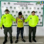 Se visualiza al capturado junto a dos uniformados de la Policía Nacional. Detrás el banner que identifica a la Policía Nacional en Tame (Arauca)