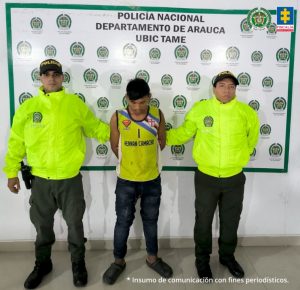 Se visualiza al capturado junto a dos uniformados de la Policía Nacional. Detrás el banner que identifica a la Policía Nacional en Tame (Arauca)