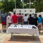 En la fotografía se encuentran diez capturados de pie, de espalda, esposados, custodiado por dos uniformados de la Policía Nacional. Delante de ellos hay una mesa con las armas, municiones y la droga incautada.