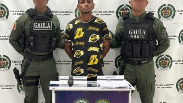 En la fotografía aparece un hombre de mediana estatura, con los brazos atrás, moreno, de cabellos crespos moreno, con barba pequeña, vestido de camiseta negra con amarillo, custodiado por dos uniformados de la Policía Nacional.