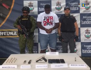 En la fotografía aparece el capturado junto a un servidor del CTI y un soldado del Gaula del Ejército. En la parte posterior está una mesa con un arma de fuego, munición, teléfonos celulares