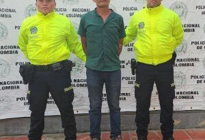 El capturado está esposado con sus manos a la espalda, viste una camisa verde y pantalón y a su lado hay dos uniformados de la Policía.