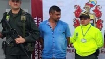 en la imagen se ve una persona detenida bajo custodia de dos uniformados de la Policía Nacional, detrás suyo un backing institucional.
