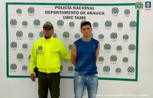 El capturado es visto junto a un uniformado de la policía nacional.  Detrás del cartel que identifica a la Policía Nacional en Tame (Arauca)