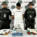 En la fotografía aparece un hombre de pie, con los brazos atrás, delgado, alto, trigueño, de cabellos negros, vestido con camiseta y pantalón blanco, custodiado por dos agentes de la Policía Nacional. Delante de ellos hay una mesa con la ropa incautada.