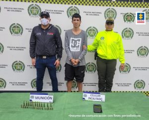 En la fotografía se aprecia de pie, con los brazos adelante y esposado a Cristhian David Zapata. Viste buso de manga larga de color gris con estampado blanco a la altura del pecho y bermuda negra. Al lado derecho de él hay un servidor del CTI de la Fiscalía y a la derecha un uniformado de la Policía Nacional. Delante de ellos, sobre una superficie verde hay munición y un teléfono celular. Asimismo, hay dos letreros que dicen: ‘Munición’. Y ‘Celular incautado’. Detrás de ellos hay un pendón institucional de la Policía.