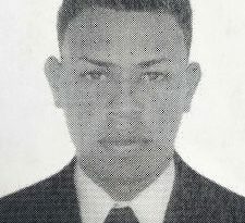 En la imagen a blanco y negro aparece el rostro de un hombre joven de ojos rasgados, nariz, boca y orejas grandes, cabellos crespos cortos, vestido con saco negro, camisa blanca y corbata negra.