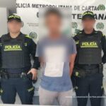 En la fotografía aparece un hombre de pie, con los brazos atrás, el rostro difuso, vestido de camiseta gris con pantalón beige, custodiado por dos agentes de la Policía Nacional. Detrás de ellos hay un pendón de esa institución.