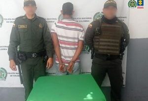 En la fotografía aparece un hombre de espalda, con los brazos esposados, vestido de camiseta blanca con rallas rojas, pantalón de jean azul, custodiado por dos agentes de la Policía Nacional.