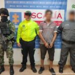 El procesado se encuentra esposado y en custodia por un servidor del Cuerpo Técnico de Investigación (CTI) Policía y Ejército Nacional. Detrás de él hay un backing de la Fiscalía.