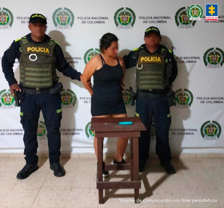 En la imagen está una mujer trigueña, se encuentra de pie, con los brazos atrás esposados, tiene un vestido ajustado negro con tenis del mismo color, cabellos recogidos negros y está custodiada por dos agentes de la Policía Nacional.