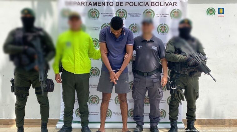 En la imagen está un hombre de pie, alto, delgado, moreno, con los brazos adelante esposado, cabellos negros, vestido de bermuda gris con camiseta azul oscura, custodiado por servidores del CTI y de la Policía Nacional.