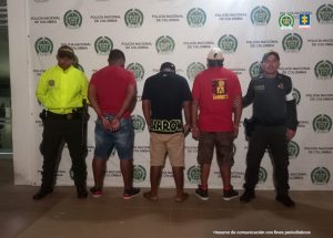 En la foto están tres hombres de pie, de espalda, vestidos con camisetas y bermudas, custodiado por dos uniformados de la Policía Nacional. Detrás de ellos hay un pendón de esa institución.