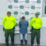 en la imagen se ve un hombre detenido bajo custodia de dos integrantes de la Policía Nacional. Detrás suyo un backing de la Policía.