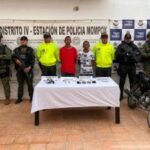 En la imagen están dos hombres de pie, ambos morenos, delgados, con los brazos atrás, acompañados por cinco uniformados de la Policía Nacional. Delante de ellos aparece una mesa con el arma, las municiones y el explosivo incautado.