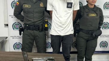 En la imagen se puede ver a un hombre detenido bajo custodia de dos miembros de la Policía Nacional.  Frente a él hay una mesa con una pistola.  Detrás del apoyo institucional.