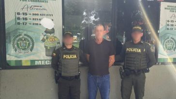 En la fotografía, aparece un hombre de pie, delgado, trigueño, con los brazos atrás esposado, vestido de camiseta negra y jean azul, custodiado por dos agentes de la Policía Nacional. Se encuentran en un CAI.