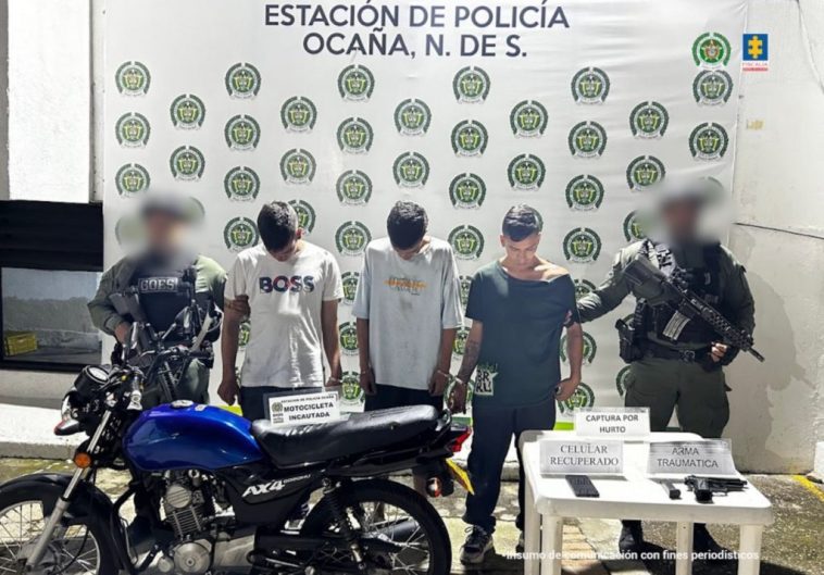 Se observa a los capturados junto a dos uniformados de la Policía Nacional. Frente a ellos una mesa con los elementos incautados durante el procedimiento de captura: celular, arma traumática y una motocicleta.