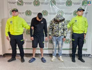 Los imputados se encuentran en custodia por dos uniformados de la Policía Nacional. Uno de los capturados tiene una pantaloneta y chaqueta negra, mientras que el otro tiene un jean con una chaqueta con capucha.  Detrás se observa un backing de la Policía.