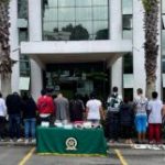 Se observan 12 personas capturadas en fila y todos de espaldas.  A cada lado de la fila, tres uniformados de la Policía Nacional. Al frente una mesa con mantel verde y logotipo de la Policía en la cual se ubican elementos incautados en las diligencias de allanamiento .