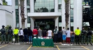 Se observan 12 personas capturadas en fila y todos de espaldas.  A cada lado de la fila, tres uniformados de la Policía Nacional. Al frente una mesa con mantel verde y logotipo de la Policía en la cual se ubican elementos incautados en las diligencias de allanamiento .