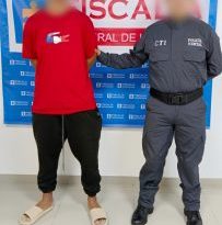 En la fotografía aparece un hombre alto, joven, de pie con los brazos atrás, trigueño, de contextura gruesa, con cabellos negros, vestido con camiseta roja, sudadera negra y chanclas beige. Está custodiado por un uniformado del CTI. Detrás de ellos hay una bandera azul de la Fiscalía General.